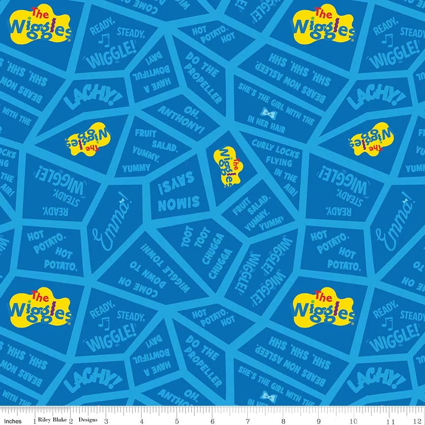 Blue fabric with a repeating The Wiggles pattern featuring yellow logo patches, white text, and darker blue geometric shapes, shown with a small ruler strip along the bottom edge.