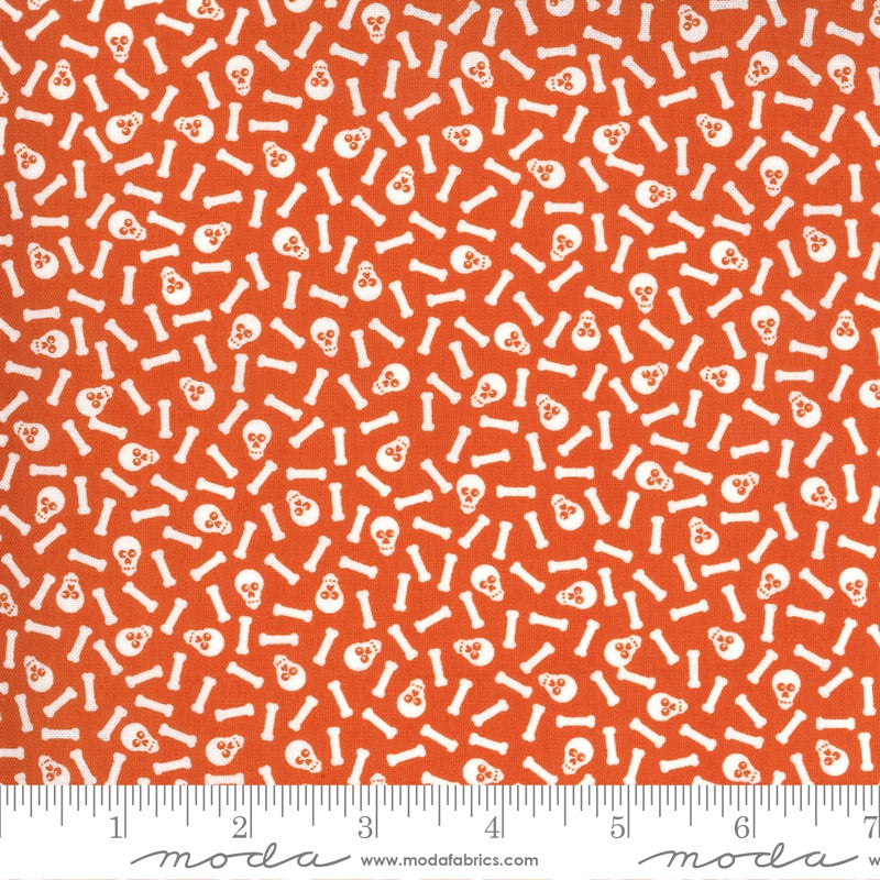 Orange fabric with a repeating white pattern of skulls and bones, shown flat with a measuring ruler along the bottom edge.