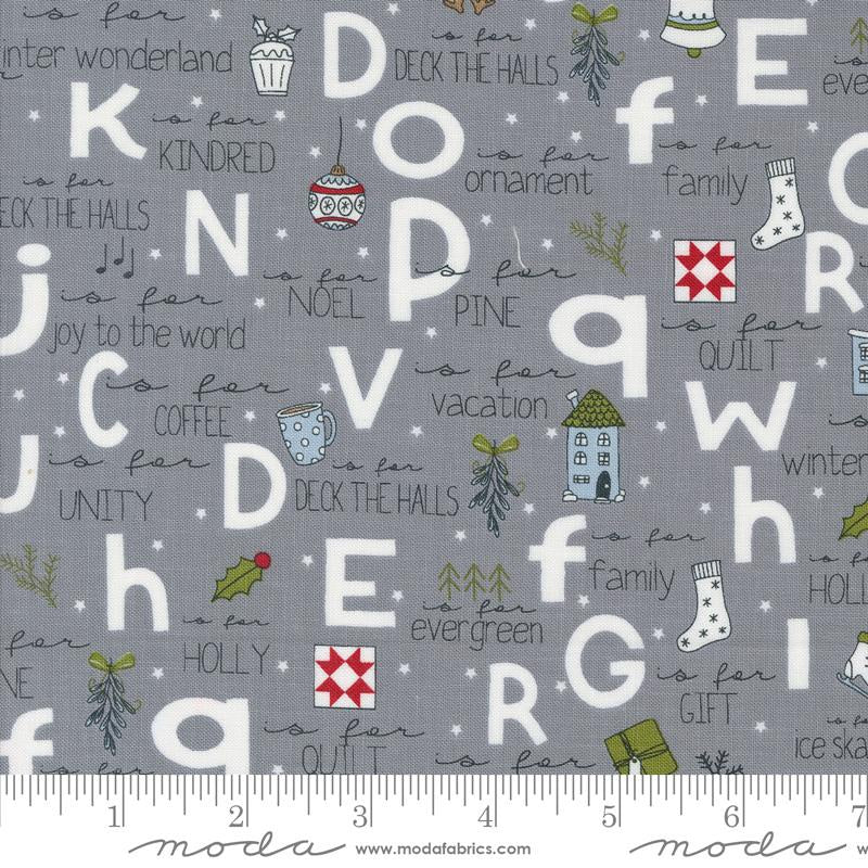 Gray fabric with a festive alphabet print featuring white letters, Christmas-themed illustrations, and phrases like 'is for family' and 'is for holiday'. A ruler along the bottom edge shows the scale of the fabric pattern.