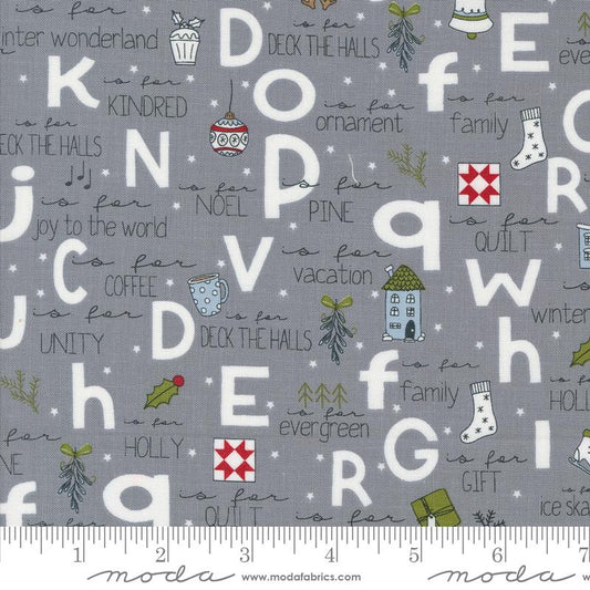 Gray fabric with a festive alphabet print featuring white letters, Christmas-themed illustrations, and phrases like 'is for family' and 'is for holiday'. A ruler along the bottom edge shows the scale of the fabric pattern.