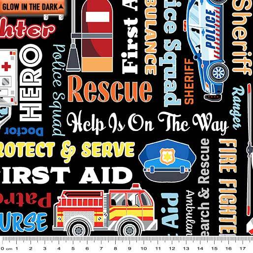 A black fabric pattern featuring various emergency services and first aid motifs, including a police car, fire truck, ambulance, police hat, and text such as 'Rescue', 'Help Is On The Way', and 'PROTECT & SERVE'. A white ruler is shown along the bottom edge to indicate scale.