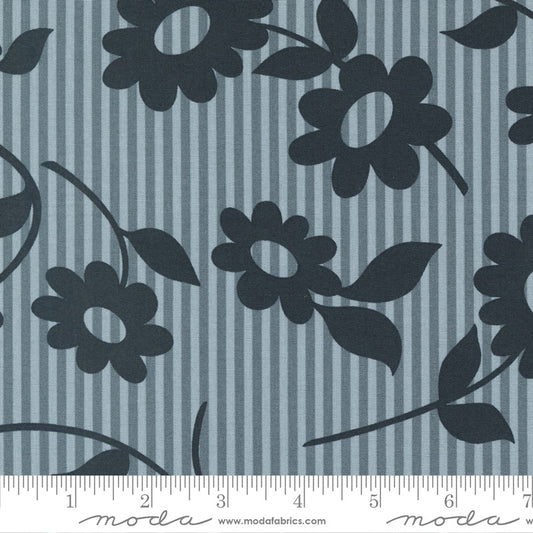 A close-up of fabric showing a repeating floral pattern with dark navy flower silhouettes and leaves over a muted blue-gray and navy vertical stripe background. A ruler along the bottom edge shows the scale.