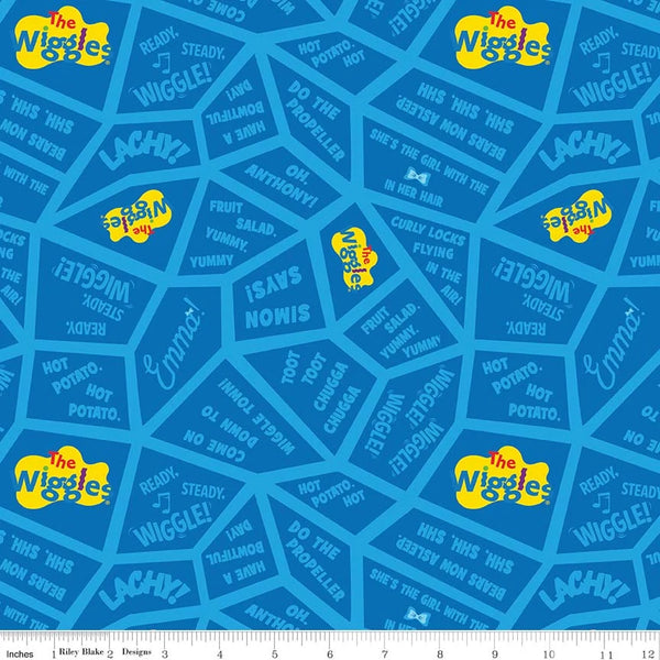 Blue fabric with a repeating The Wiggles pattern featuring yellow logo patches, white text, and darker blue geometric shapes, shown with a small ruler strip along the bottom edge.