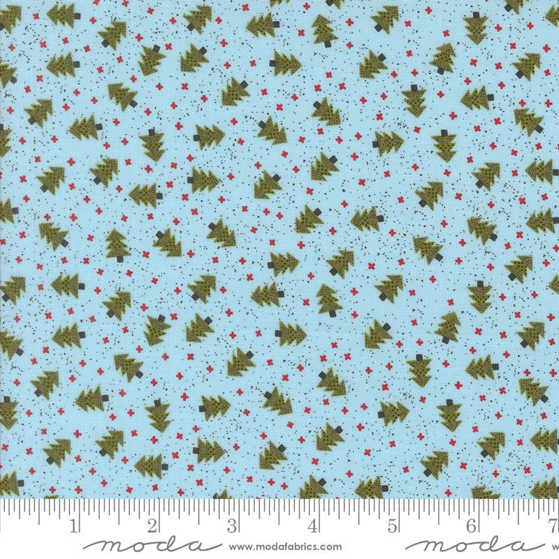 Light blue fabric with a repeating pattern of olive green Christmas trees and small red stars, shown with a ruler along the bottom edge for scale. The brand name 'Moda' is visible at the bottom.