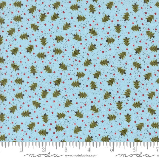 Light blue fabric with a repeating pattern of olive green Christmas trees and small red stars, shown with a ruler along the bottom edge for scale. The brand name 'Moda' is visible at the bottom.