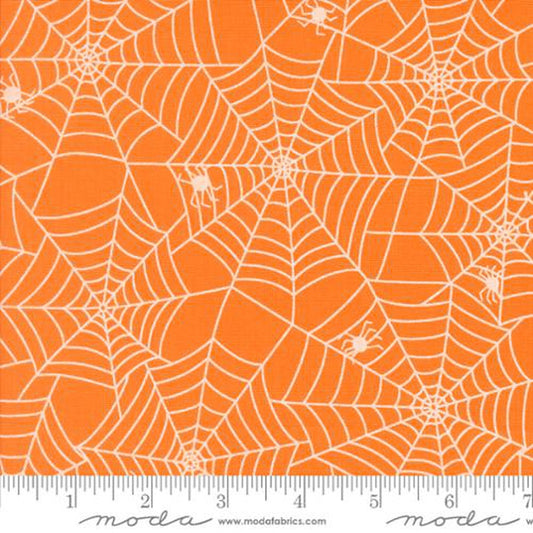 Orange fabric with a white spiderweb and spider pattern, shown flat with a ruler along the bottom edge for scale. The brand name 'Moda Fabrics' is visible on the ruler watermark.