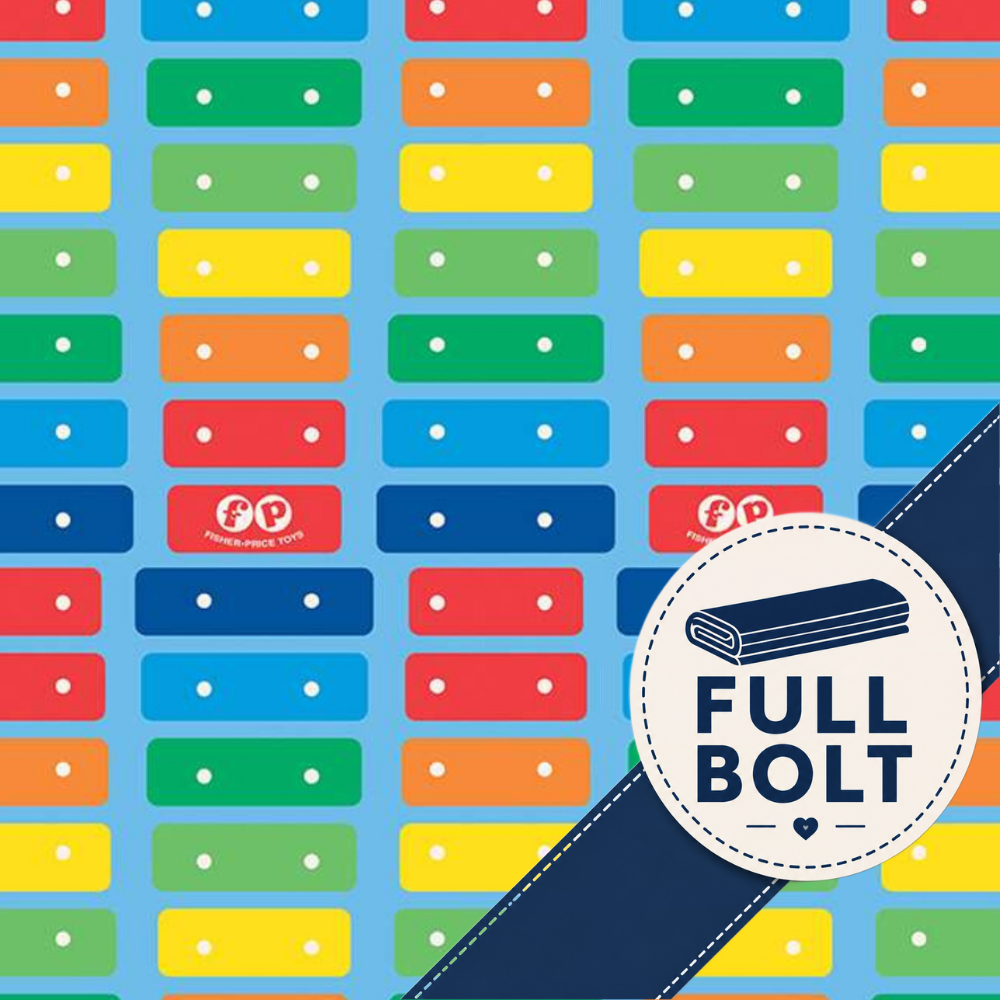 A repeating fabric pattern featuring colorful rectangular blocks in red, orange, yellow, green, and blue with small white dots, shown on a light blue background with a ruler along the bottom edge.