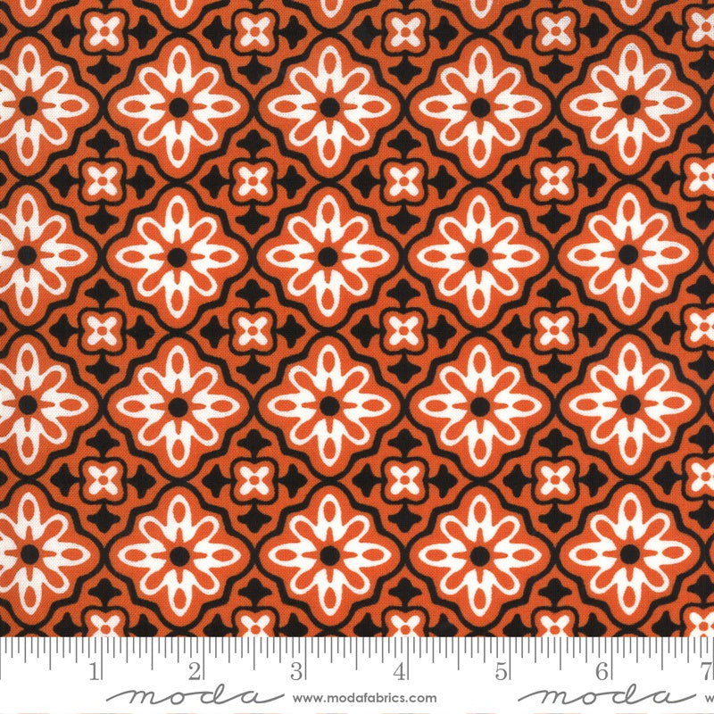A close-up of a repeating fabric pattern with orange, white, and black geometric floral motifs, shown with a ruler along the bottom edge for scale. The brand name 'moda' is visible at the bottom.