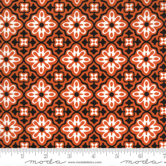 A close-up of a repeating fabric pattern with orange, white, and black geometric floral motifs, shown with a ruler along the bottom edge for scale. The brand name 'moda' is visible at the bottom.