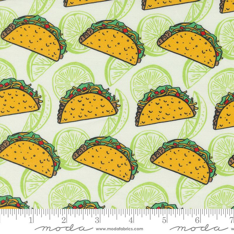 A close-up of a fabric pattern featuring yellow tacos with green lettuce and red peppers on a white background with light green lime slices. A measuring ruler is visible along the bottom edge, and the Modafabrics website watermark is printed on the ruler.