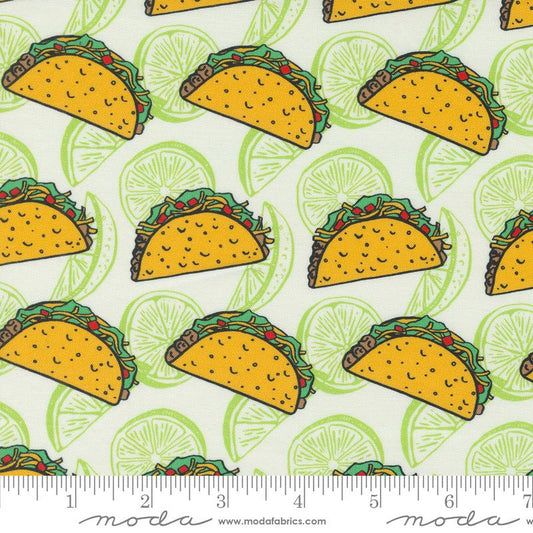 A close-up of a fabric pattern featuring yellow tacos with green lettuce and red peppers on a white background with light green lime slices. A measuring ruler is visible along the bottom edge, and the Modafabrics website watermark is printed on the ruler.