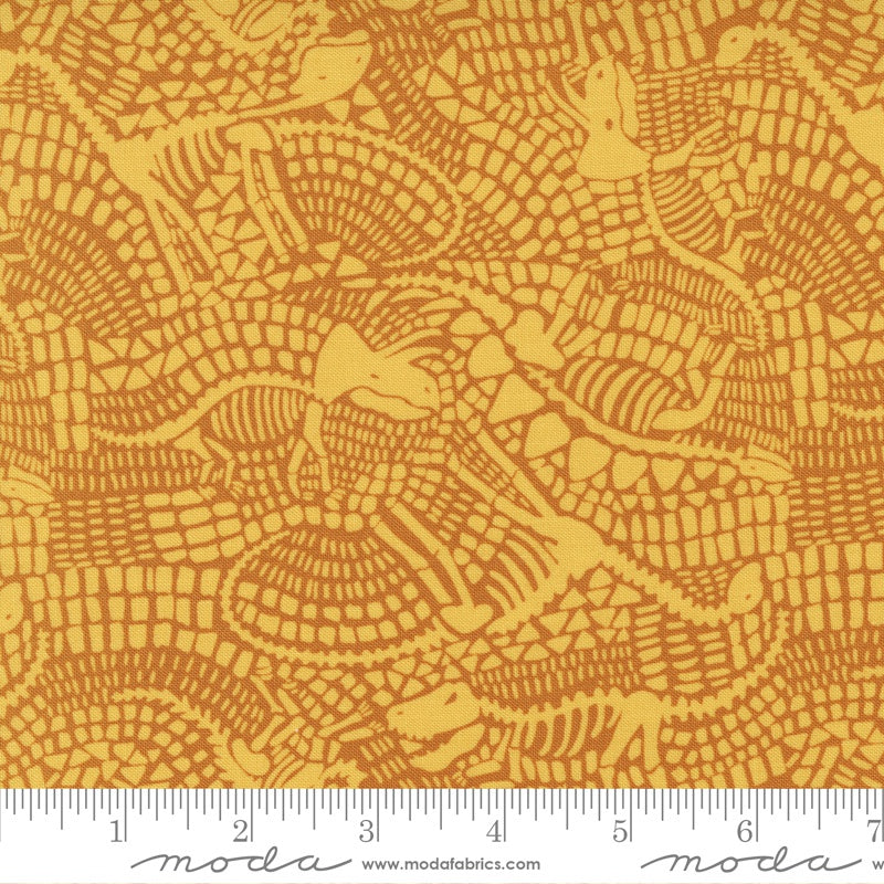 A close-up of fabric with a repeating alligator print in mustard yellow and orange-brown dinosaur fossils , with a measuring ruler along the bottom edge showing the scale of the pattern.