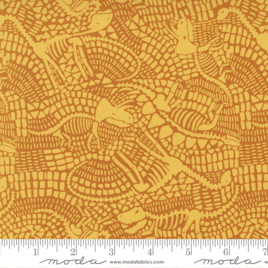 A close-up of fabric with a repeating alligator print in mustard yellow and orange-brown dinosaur fossils , with a measuring ruler along the bottom edge showing the scale of the pattern.