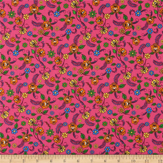 A close-up of a pink patterned fabric with a repeating floral and vine design in yellow, orange, blue, green, and purple, with a wooden ruler along the bottom edge showing measurements.