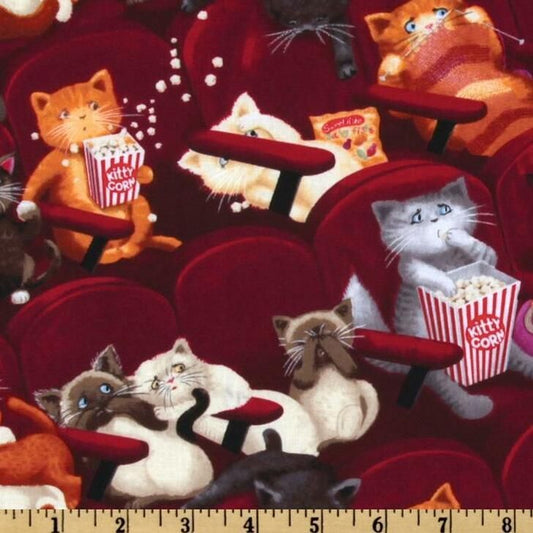 A close-up of fabric featuring a repeating pattern of cats lounging in red theater seats, each holding a striped popcorn bucket labeled 'Kitty Cool.' A wooden ruler is visible along the bottom edge for scale.